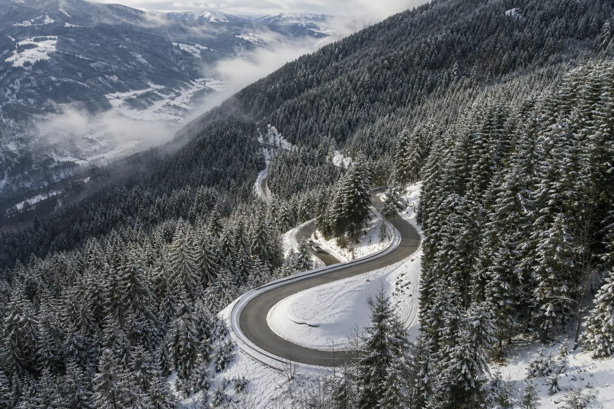 Route de montagne enneigée serpentant entre des sapins sous un ciel gris d’hiver